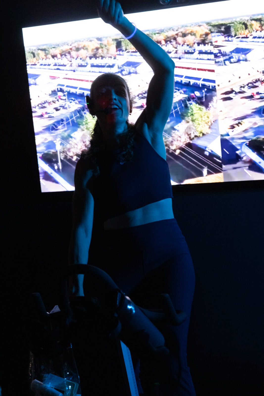 Instructor with a headset mic, arm raised, silhouetted in blue light in front of the screen