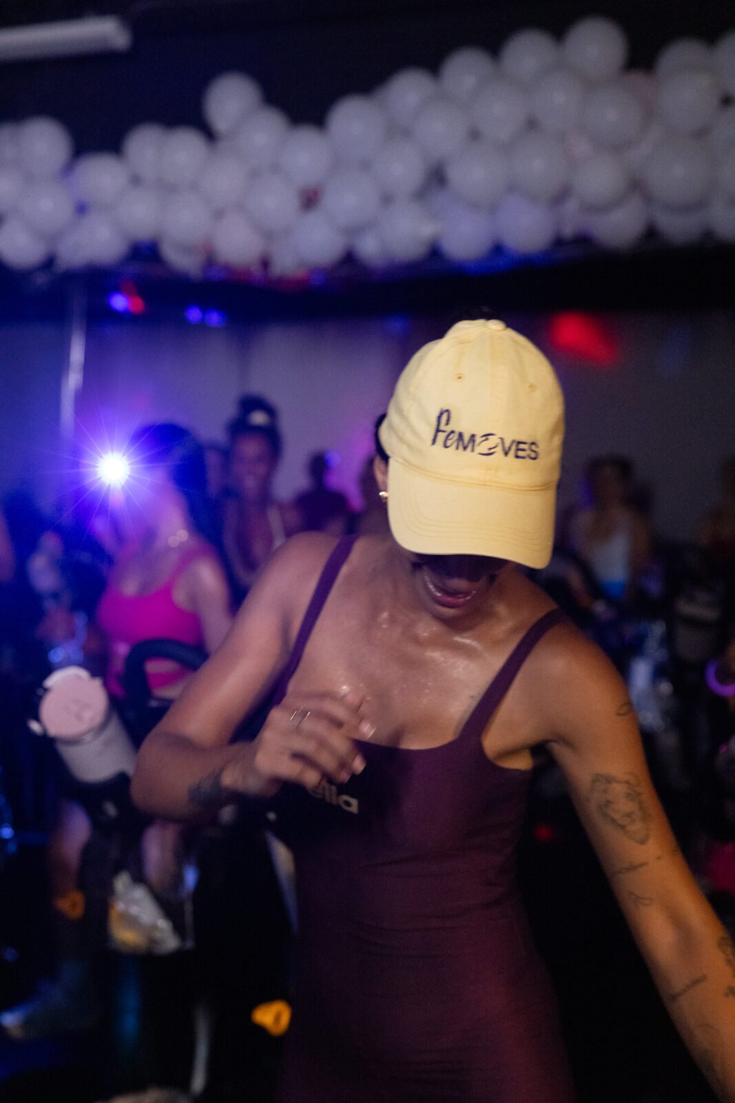 A FeMoves instructor in a plum one-piece and butter-yellow FeMoves cap dancing in the aisle